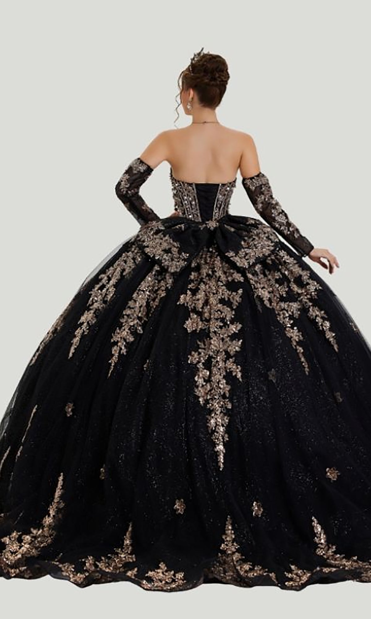 A woman stands with her back to the camera, showcasing the stunning Quinceanera Dress 26402 By Stella Couture. This strapless black ball gown features gold floral embroidery, a beaded corset bodice, and matching long gloves, while a glitter tulle skirt completes the look with an elegant flourish.